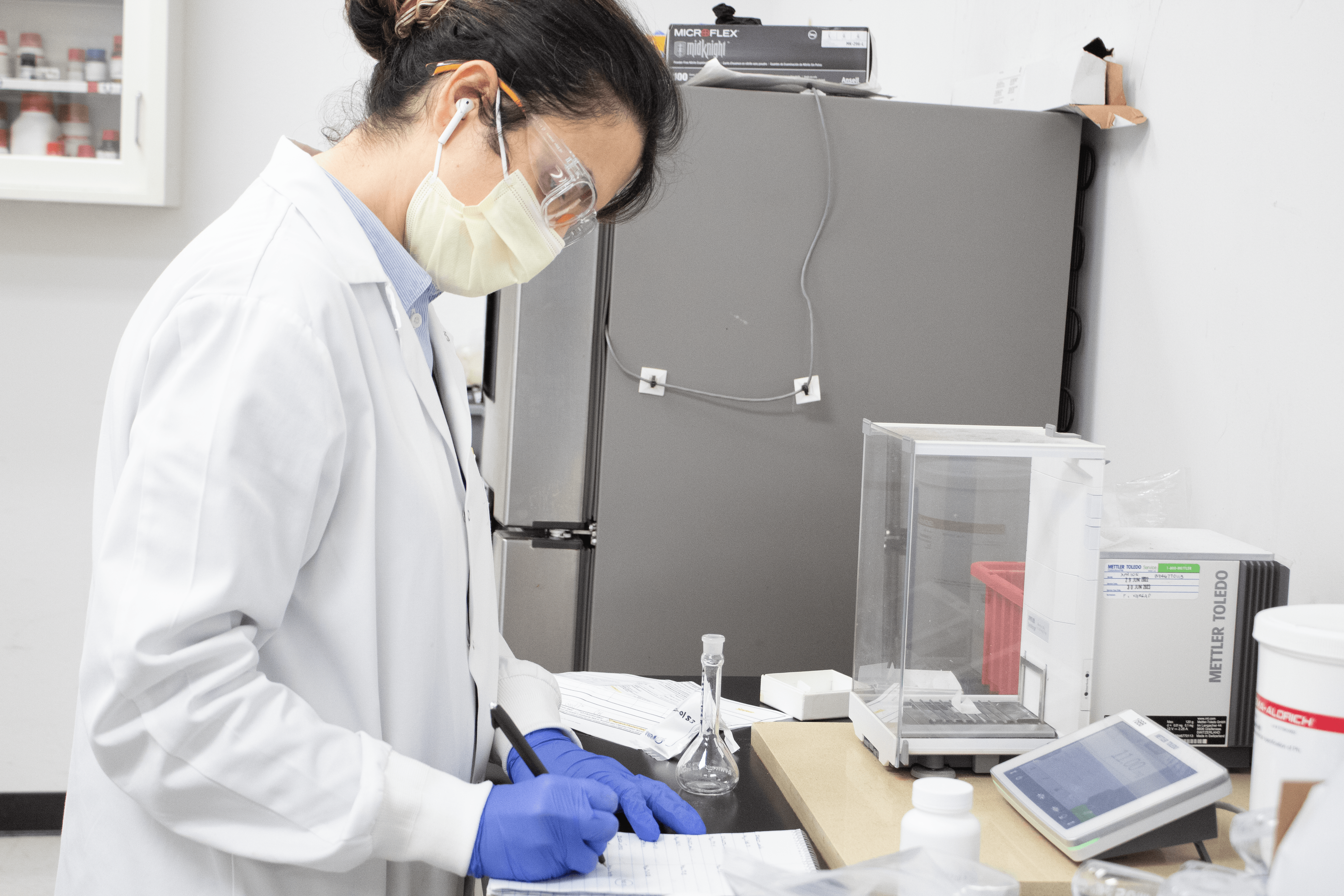 Scientist in lab wearing lab coat writing information from cosmetic stability testing on a tablet.