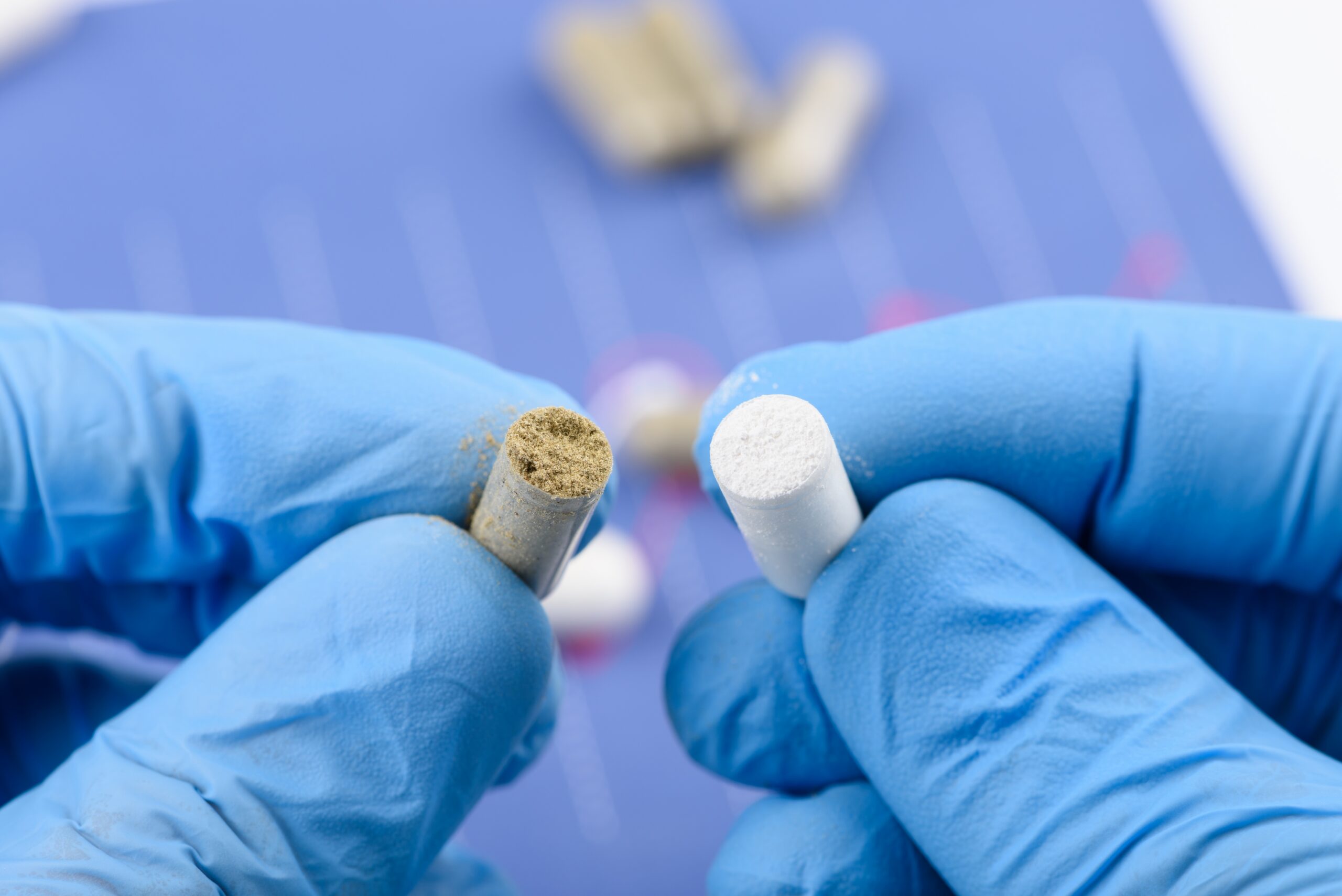 Gloved hands hold two capsules—one brown powder, one white—in a lab setting.