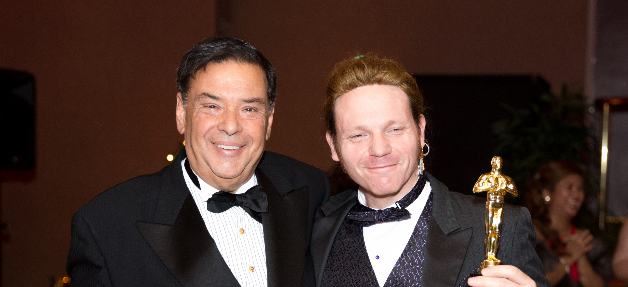 Two men in tuxedos smile for the camera at an awards event, with one holding a gold Oscar statue in the foreground.