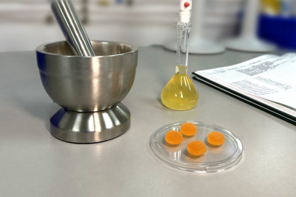 Stainless steel pestle, flask, and orange dietary supplement gummies on a table in a testing laboratory. 